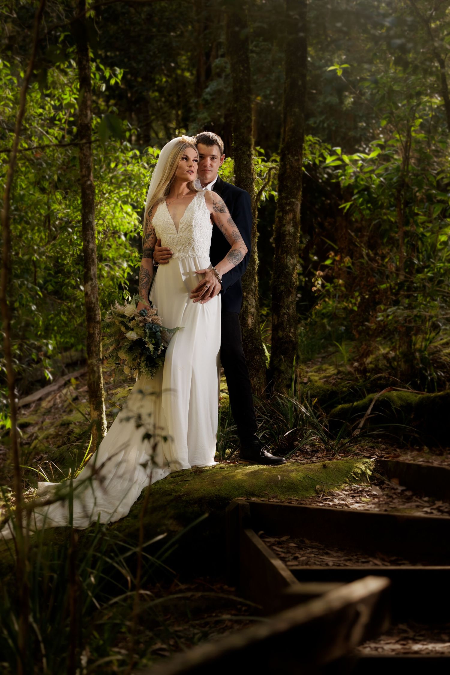 A romantic wedding couple stands in a sunlit forest setting with a white gown and formal attire.