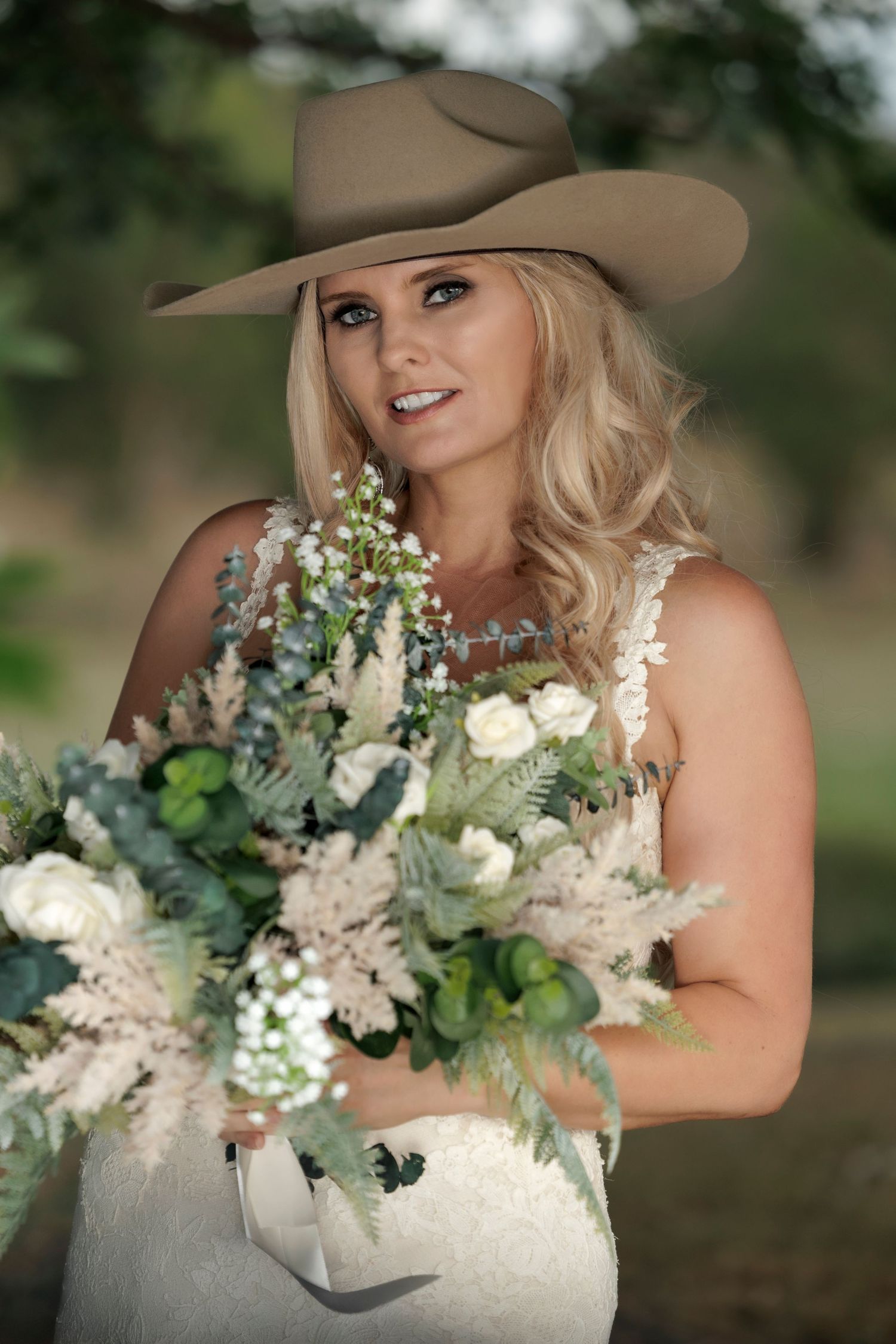 Western bride in tan cowboy hat holds lush white and green floral bouquet during outdoor country wedding photoshoot.