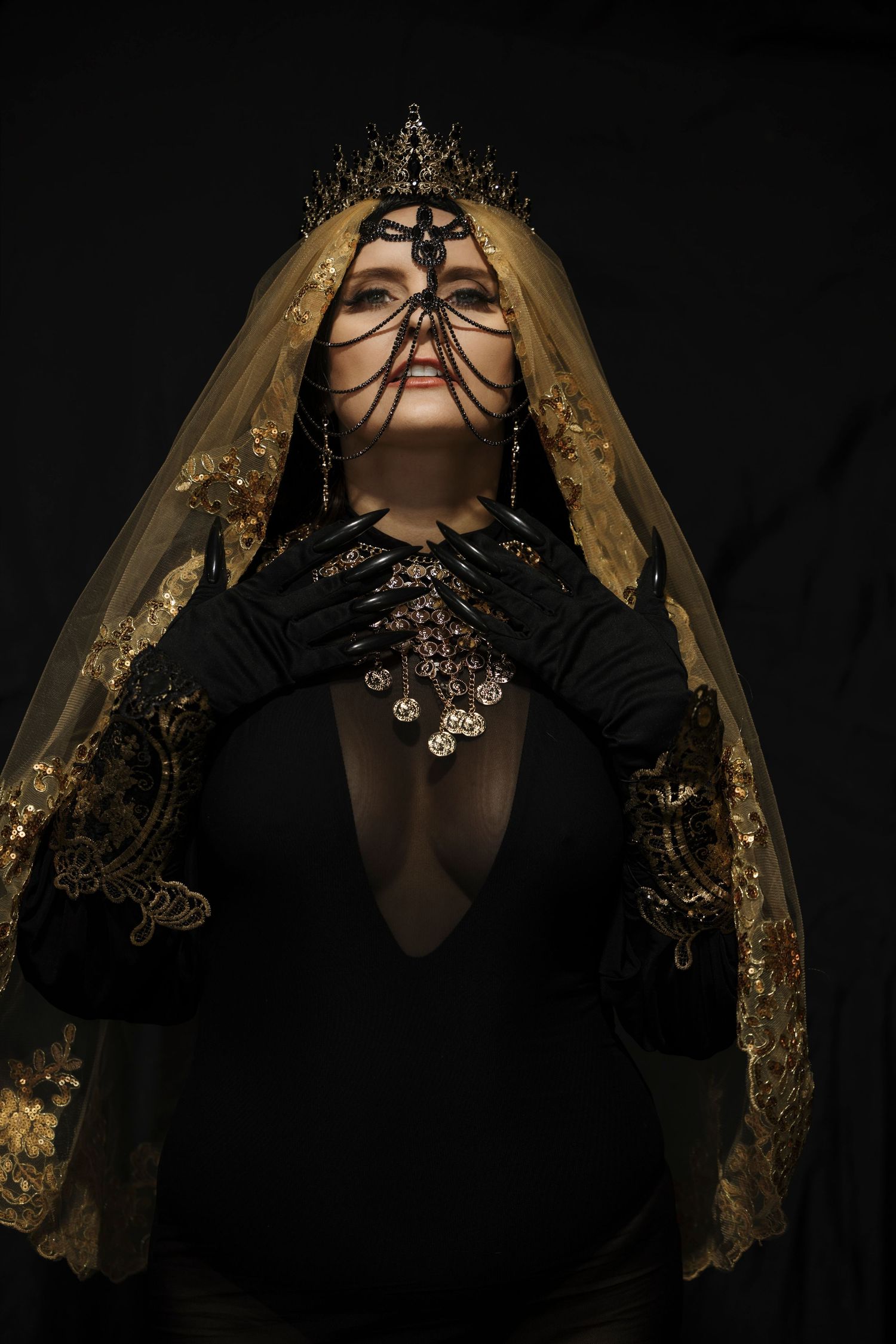 Dramatic dark portrait featuring black clothing, ornate gold veil, and antique coin jewelry against black background.