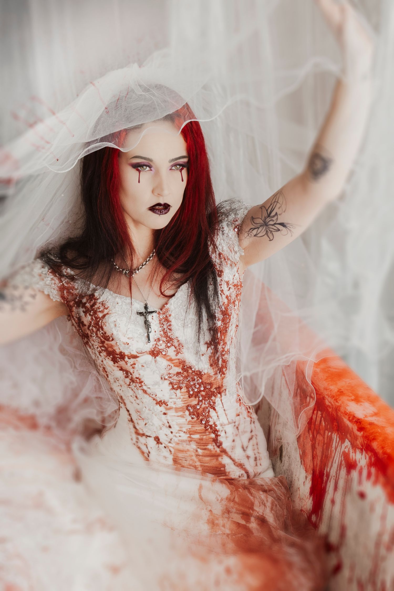 Gothic bride in lace dress with red hair holding tulle veil in ethereal setting.
