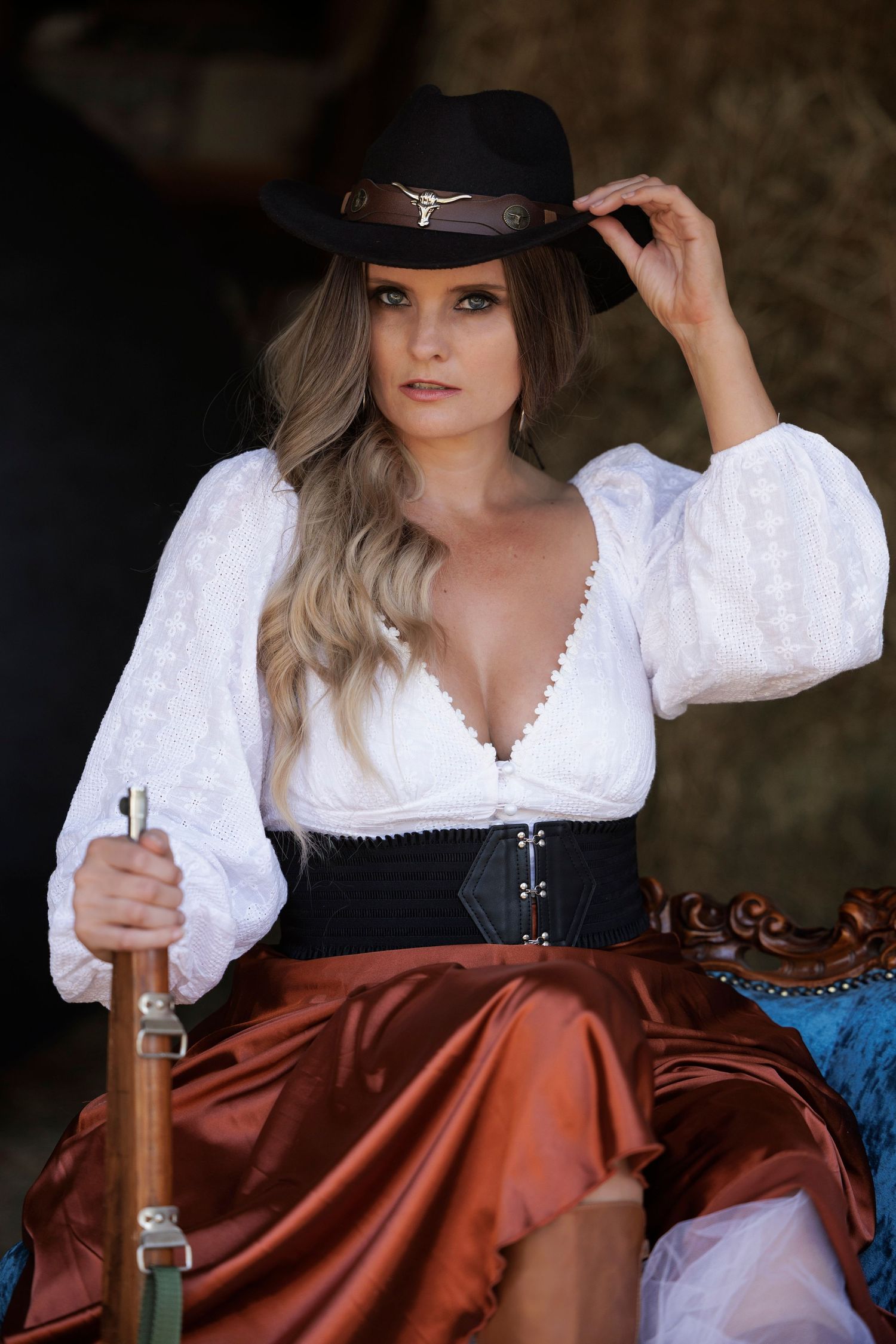 Portrait of a woman in a white blouse, black corset and brown skirt wearing a dark cowgirl hat in a rustic setting.
