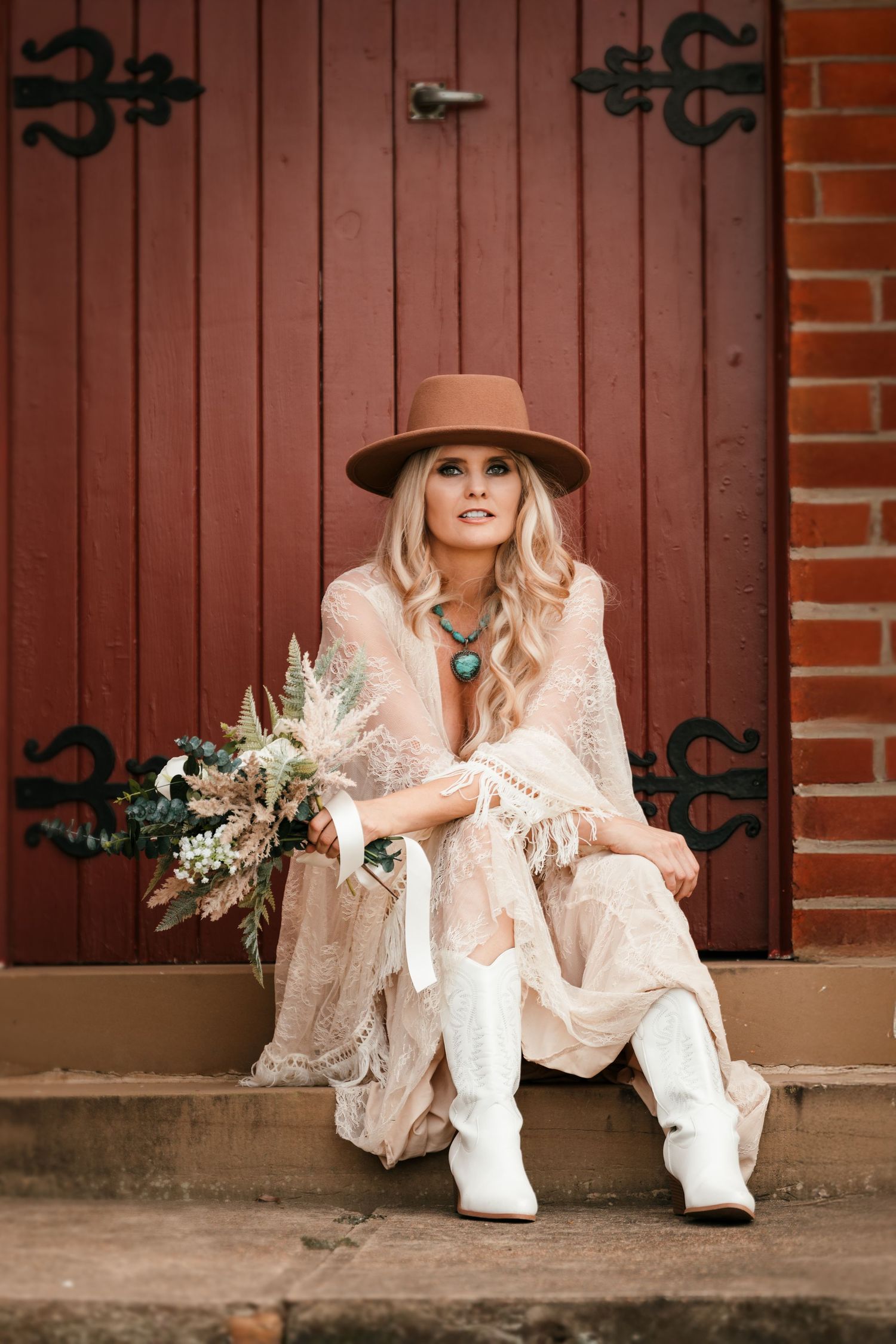 Boho-style figure in lace dress and brown hat sits on steps by rustic red door holding dried flower bouquet.