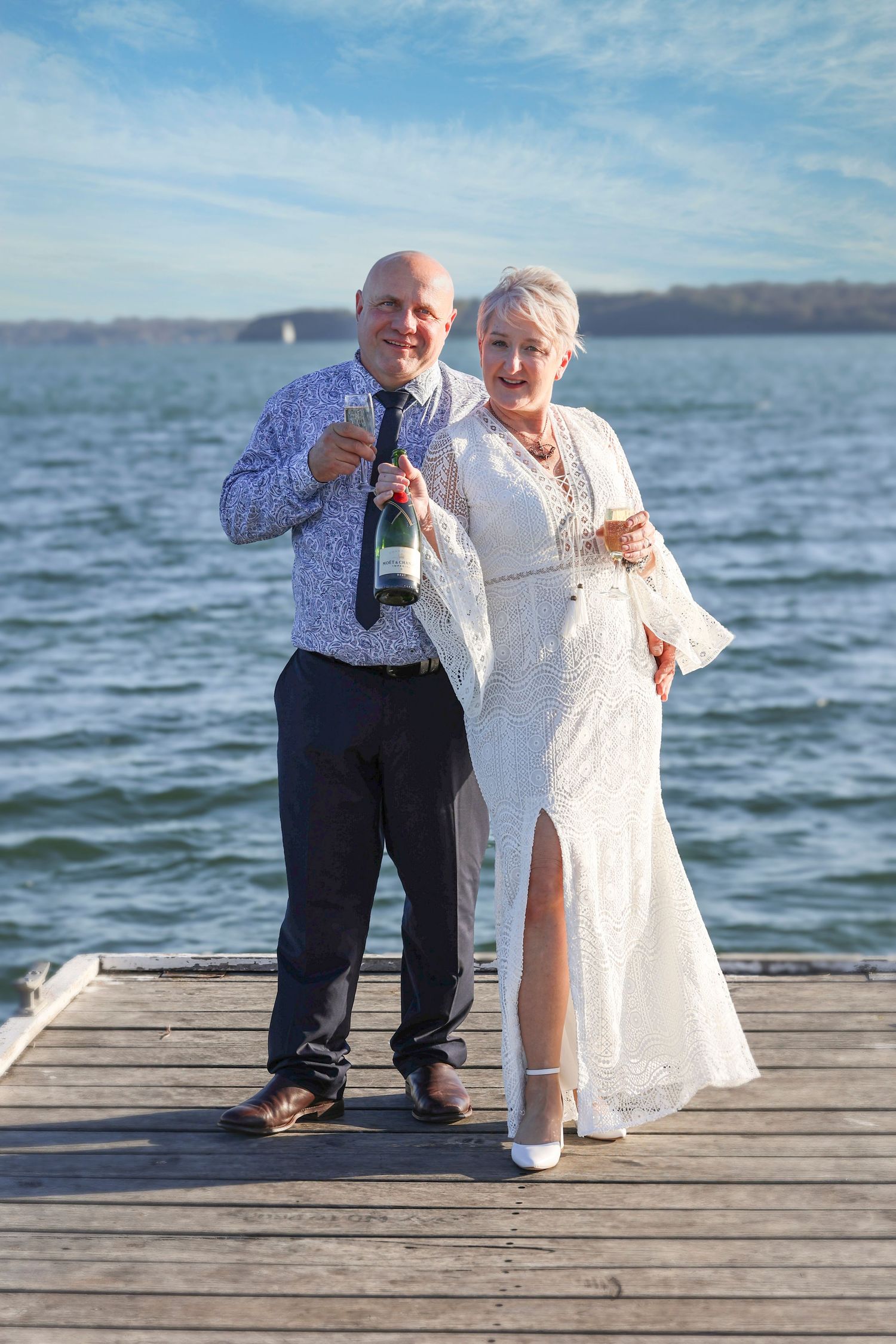 A couple celebrates on a dock overlooking a lake, holding champagne glasses on a sunny day.