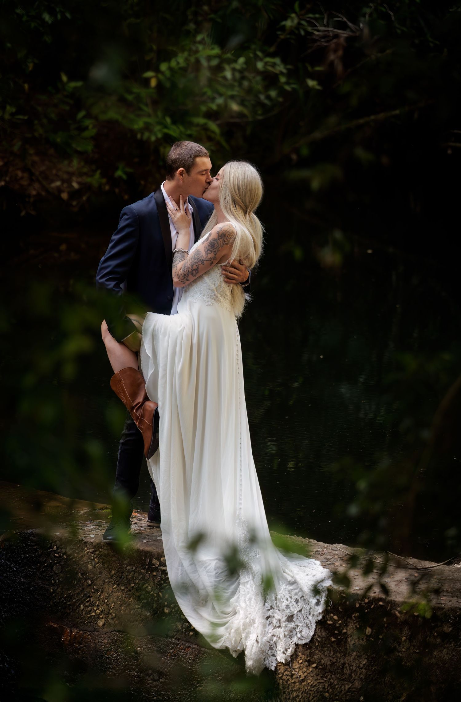 A romantic wedding couple share a tender kiss while standing on a rustic stone wall surrounded by lush greenery.