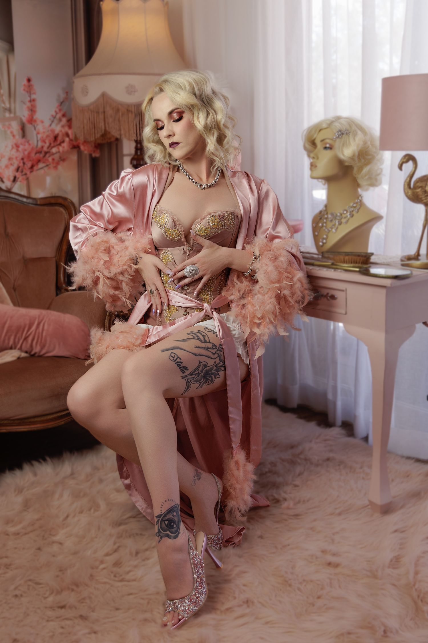 A person in a pink feathered robe and glittery shoes poses on a plush white carpet in a boudoir setting.
