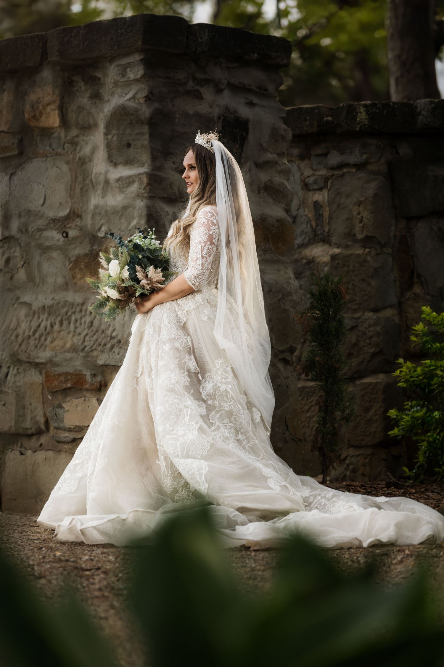An elegant bride in a lace wedding gown with flowing veil poses against a stone wall in a garden setting.