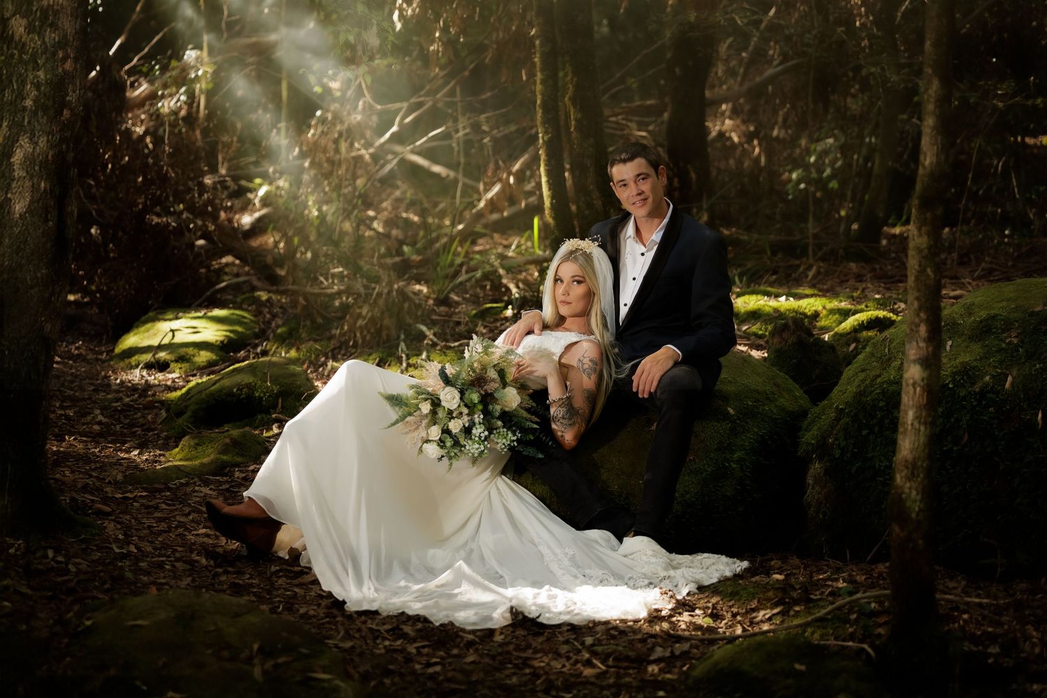 A romantic wedding couple poses in an enchanted forest setting with dramatic lighting and lush green ferns.