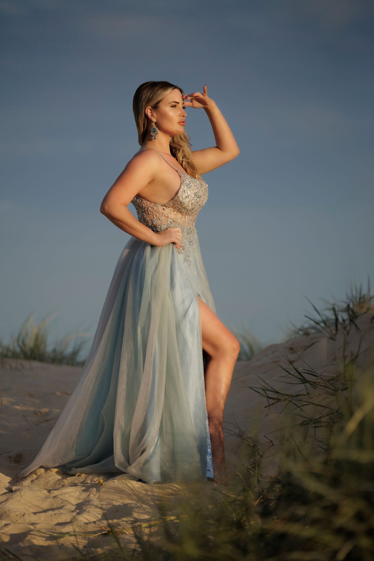 Woman in flowing blue gown poses on sand dunes at golden hour.