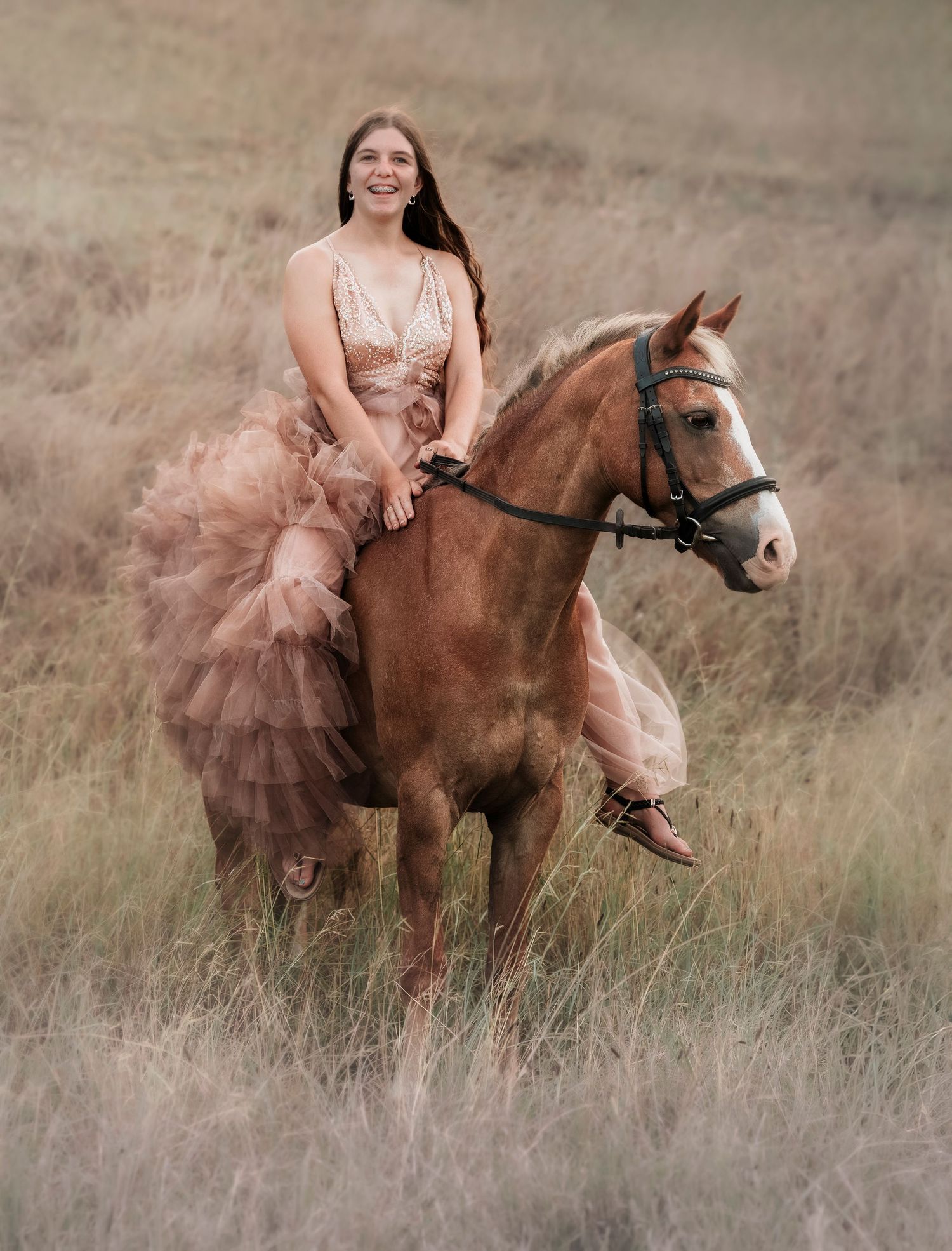 A girl with long dark hair in a flowing pink tulle dress rides a palomino horse through tall golden grass in a dreamy outdoor portrait.