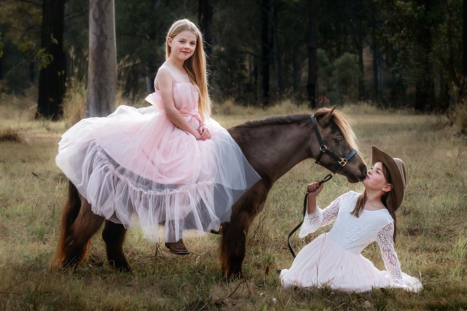 Two figures in flowing dresses pose with a brown pony in a forest setting.