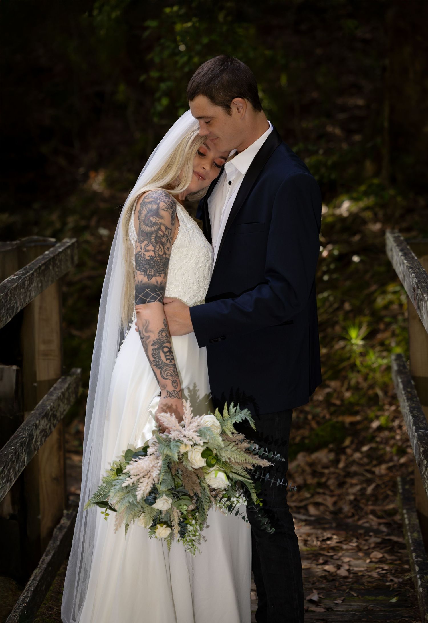 Couple shares intimate wedding moment on wooden bridge with bride holding elegant white and green bouquet.