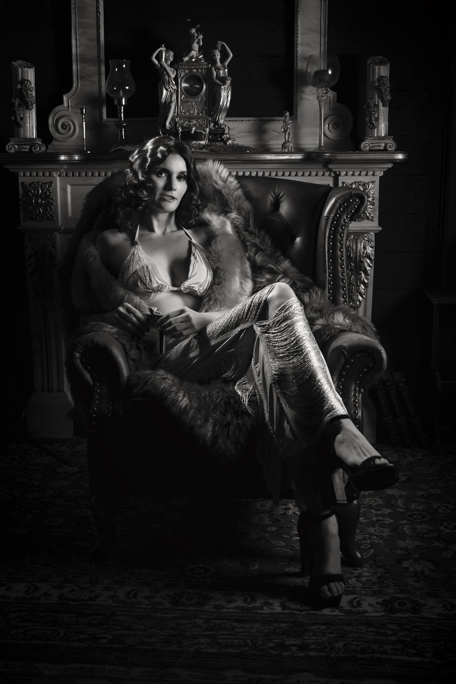Dramatic black and white portrait of a woman lounging in an ornate vintage armchair with decorative mantle in background.