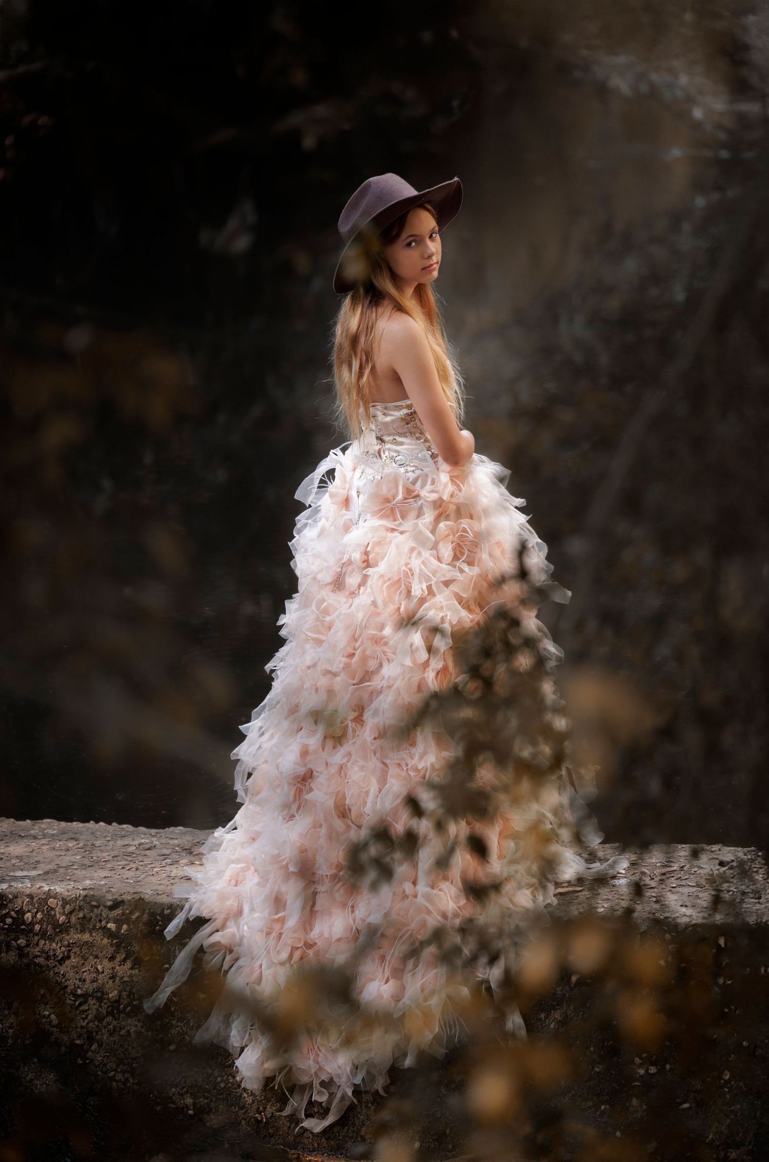 Dreamy portrait of figure in ruffled pink dress and hat against dark moody background.