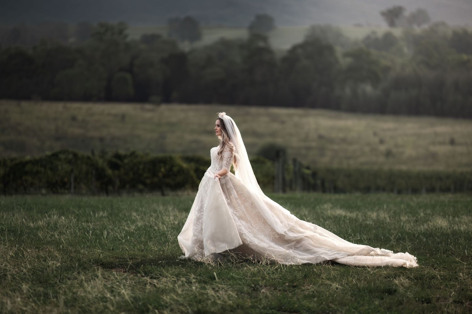 A flowing white wedding dress with long train billows in the wind across a misty green meadow at dusk.