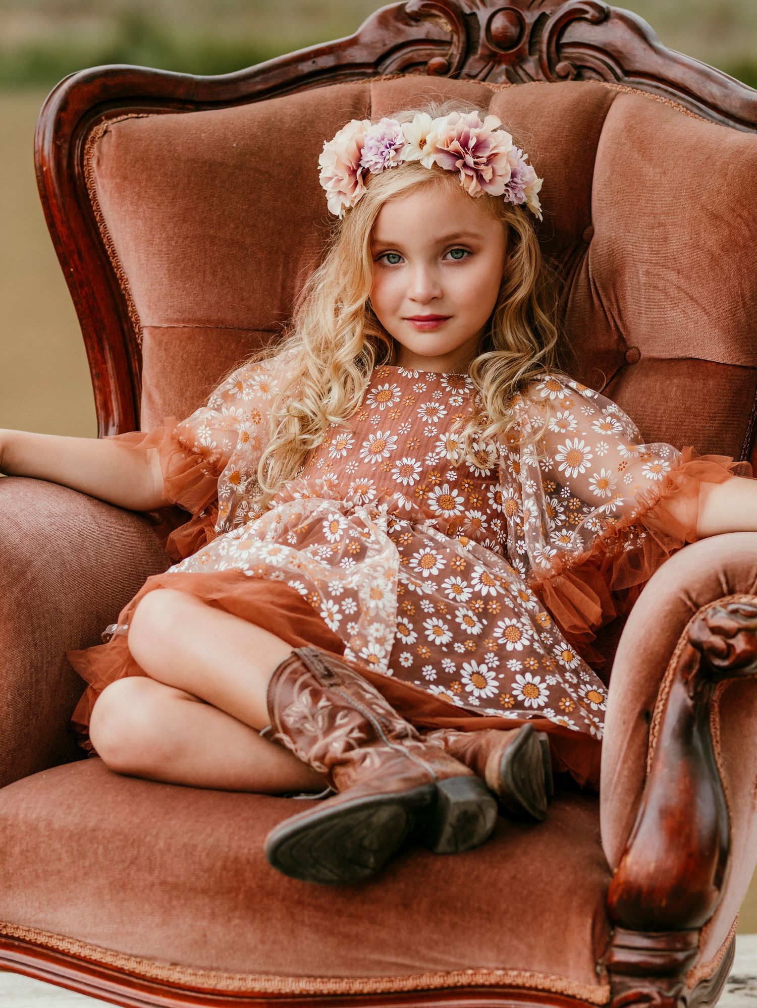 Child in vintage floral dress and flower crown poses on antique velvet chair.