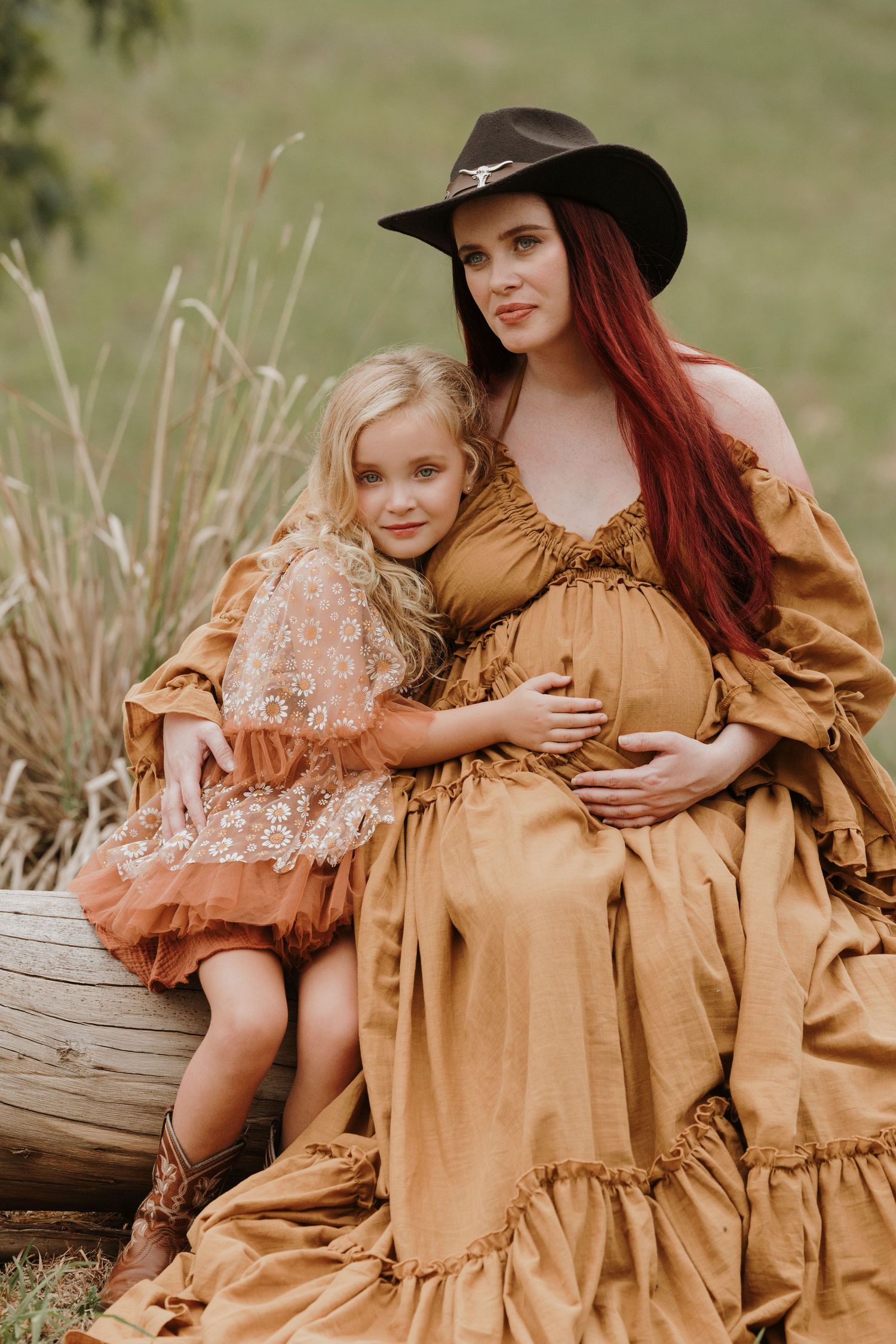 A rustic outdoor scene with a mother and daughter in vintage-inspired brown dresses sitting on a fallen log.