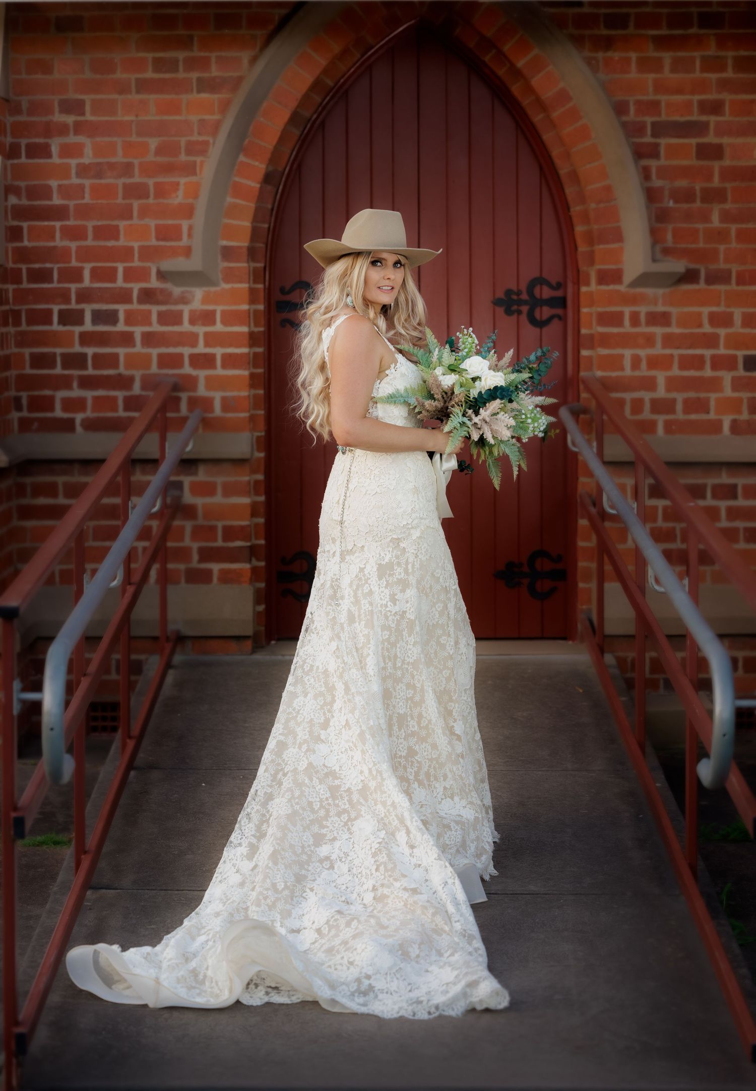 Bride in a lace wedding dress and cowboy hat stands in front of an arched brick church doorway holding a bouquet.