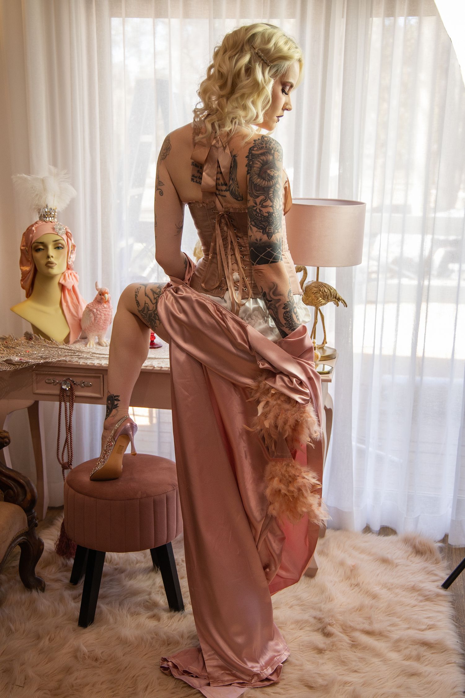 A figure in a pink dress poses on a white fluffy rug in a softly lit room with sheer curtains in the background.