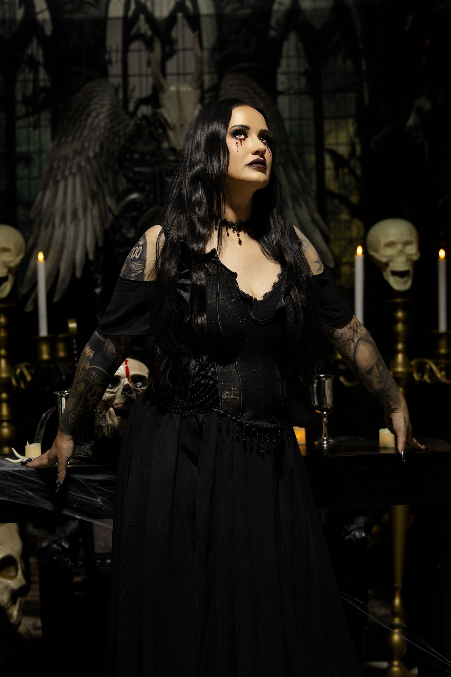 A dark gothic figure in black lace dress poses dramatically with candles and angel wings in a moody setting.