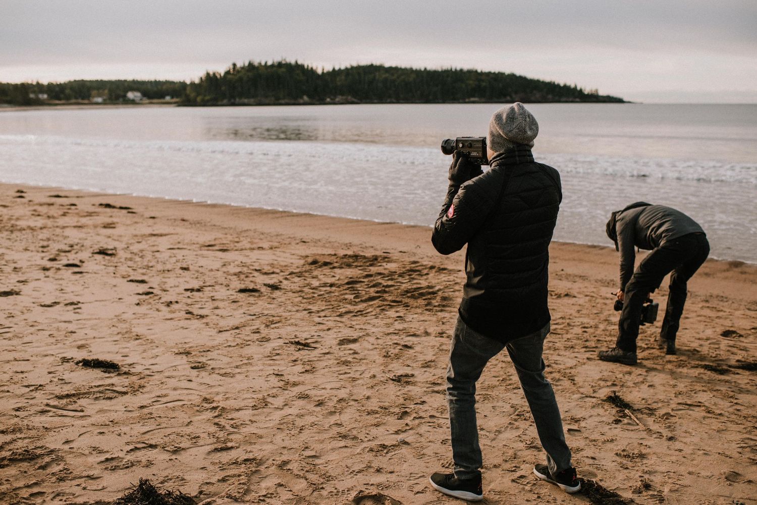 Two photographers work on a sandy beach with calm waters and forested hills in the background on an overcast day.