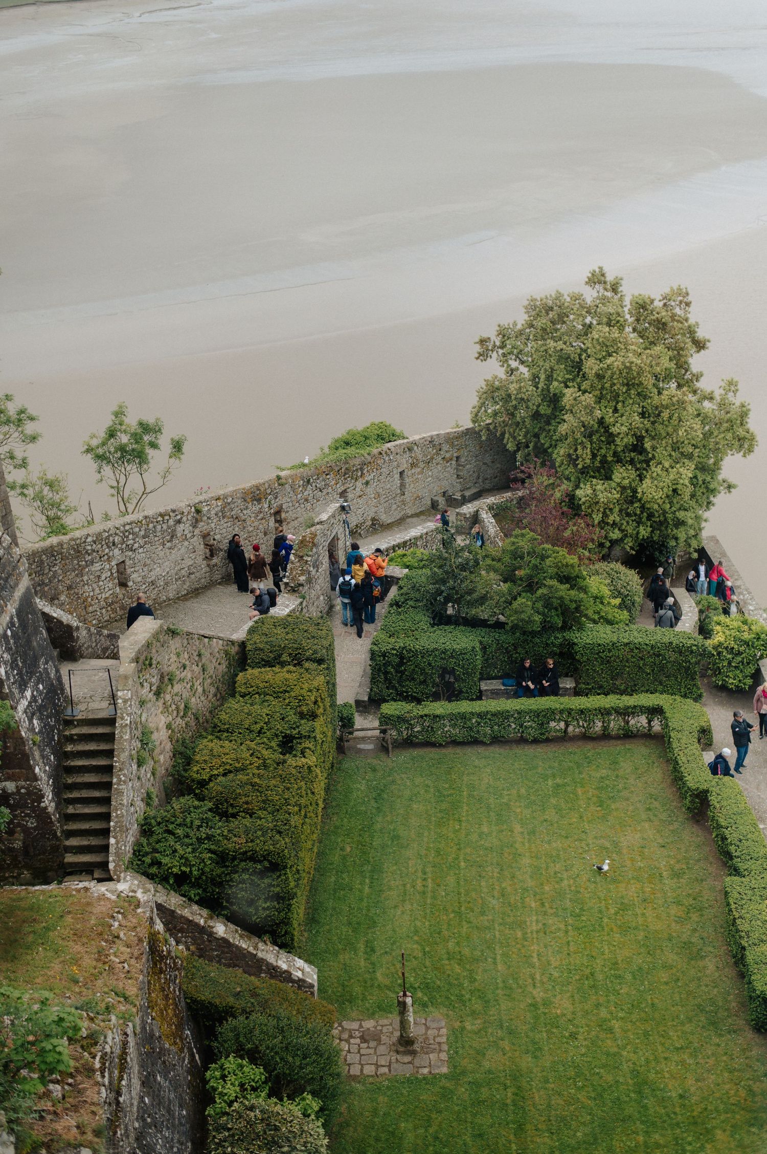 Aerial view of a historic stone garden with manicured hedges and tourists walking along a coastal wall.