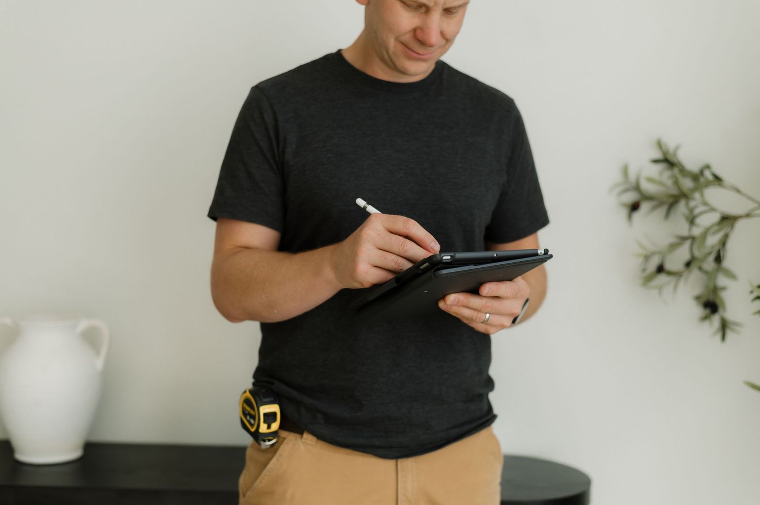 A man in a black t-shirt uses a stylus on a tablet while wearing a tool belt around his waist.