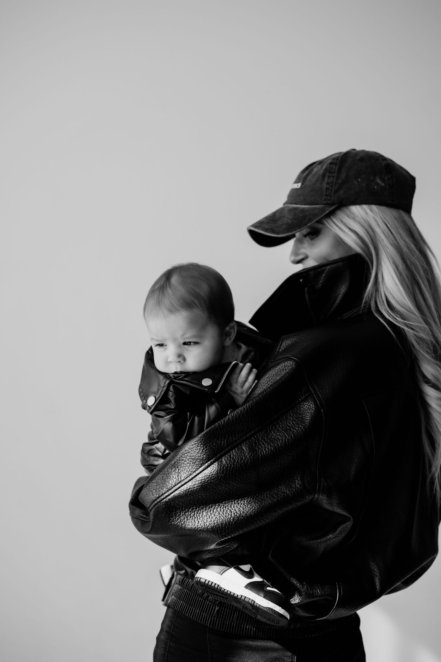 Black and white photo of a blonde woman in a leather jacket and cap tenderly holding a baby.