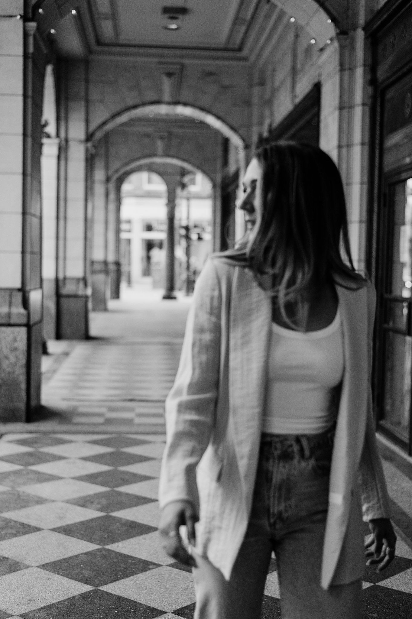 Black and white photo of a woman in jeans and blazer standing in an elegant archway corridor with checkered floors.