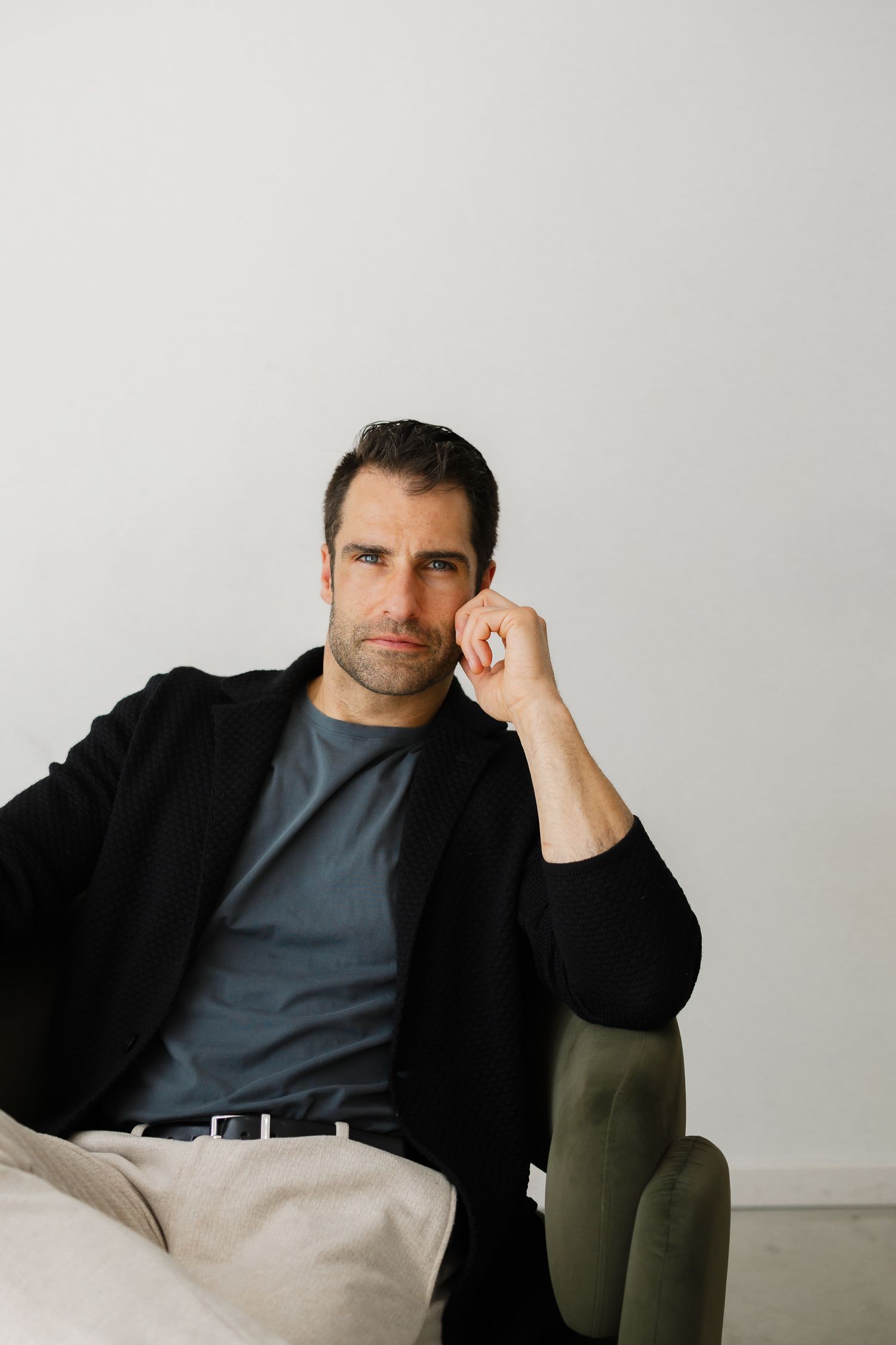A man in a black blazer and gray t-shirt sits relaxed in a green chair, resting his hand near his face.