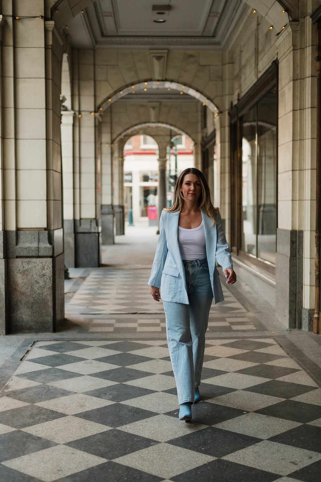 Woman in light blue blazer and jeans walks through elegant arched colonnade with checkered marble floor.