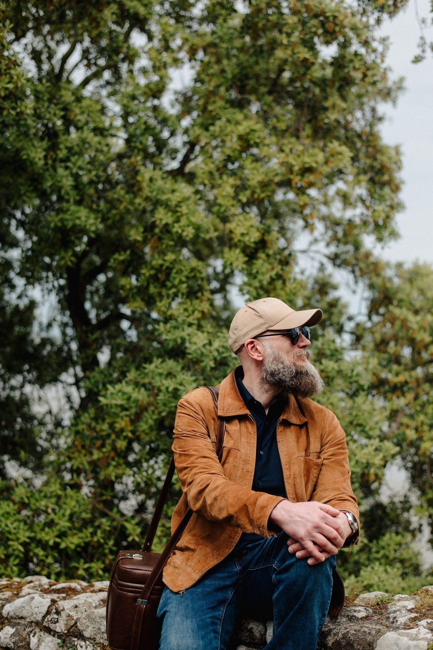 Bearded man in a tan cap and brown jacket sitting on a stone wall surrounded by lush green trees.