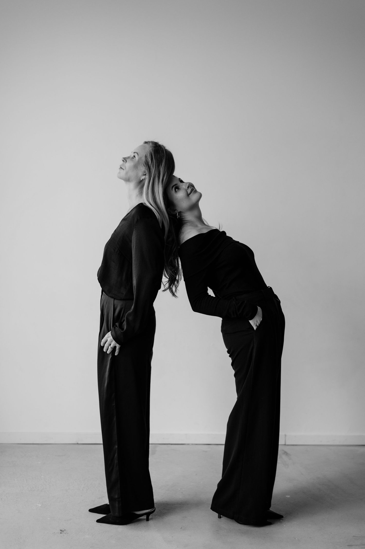 Two women in black outfits pose back-to-back, leaning heads back elegantly in a black and white studio photo.