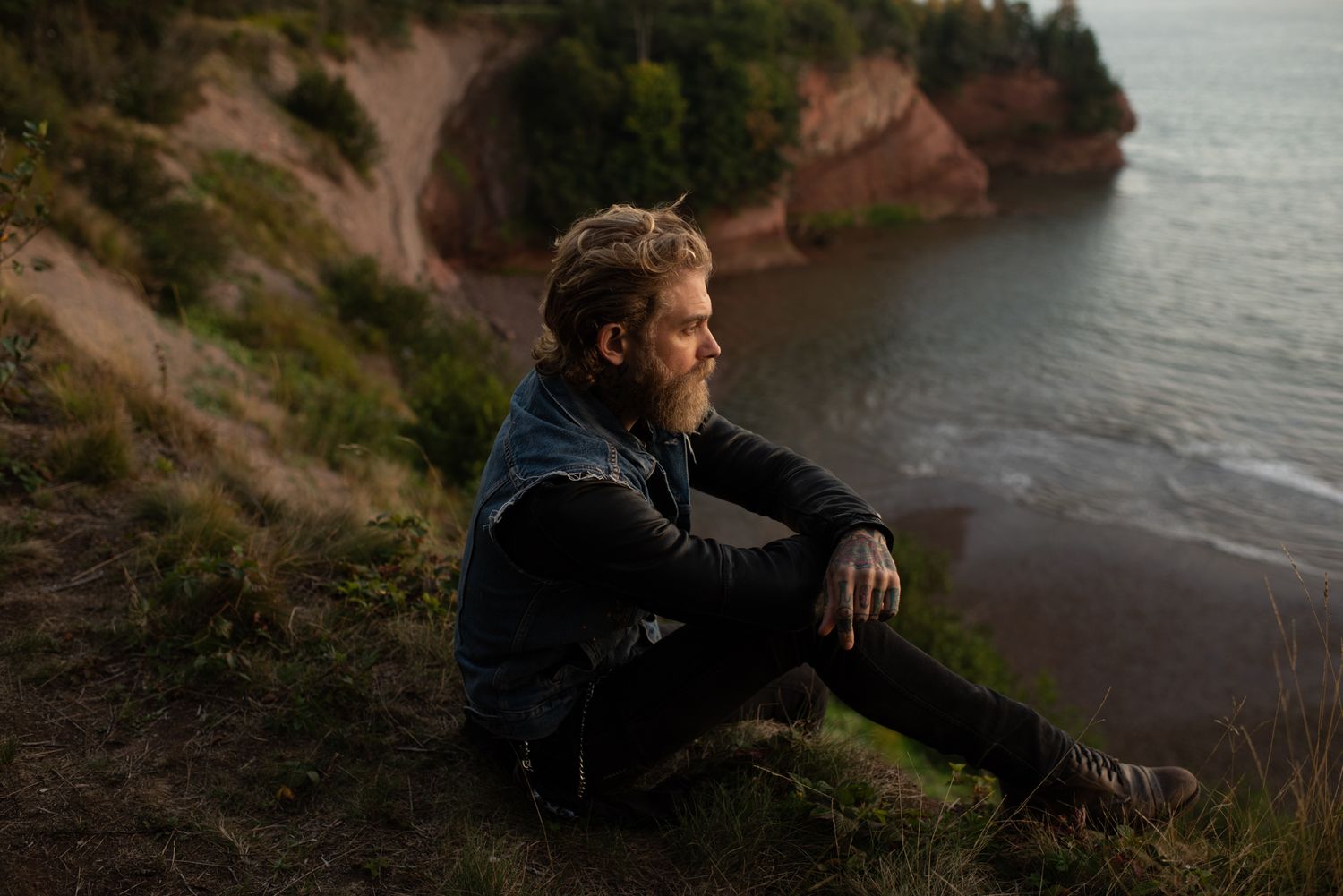 Bearded man sitting on a coastal cliff edge gazing at the ocean at dusk.