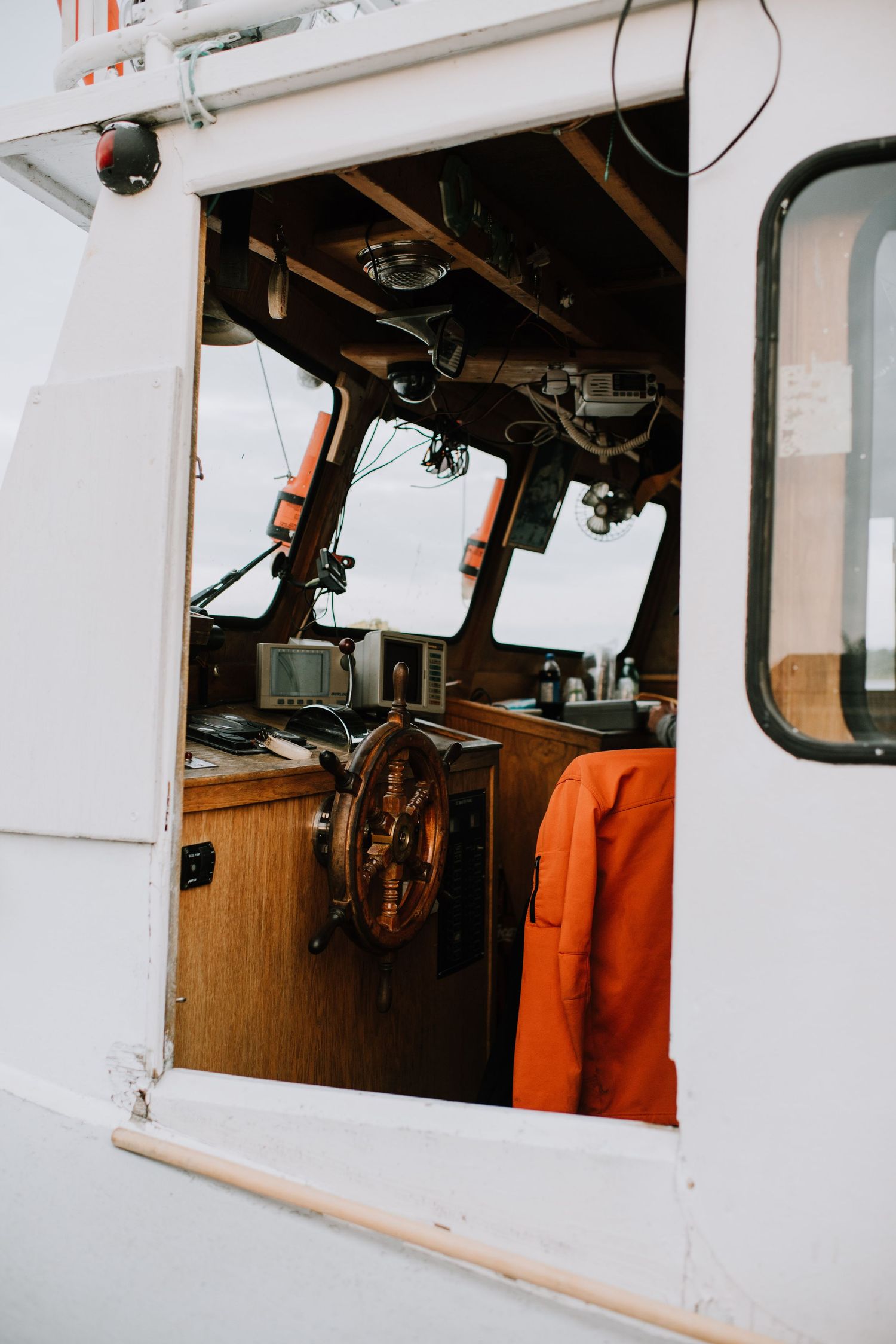 View inside a boat wheelhouse with wooden helm, navigation equipment, and orange jacket visible through open door.