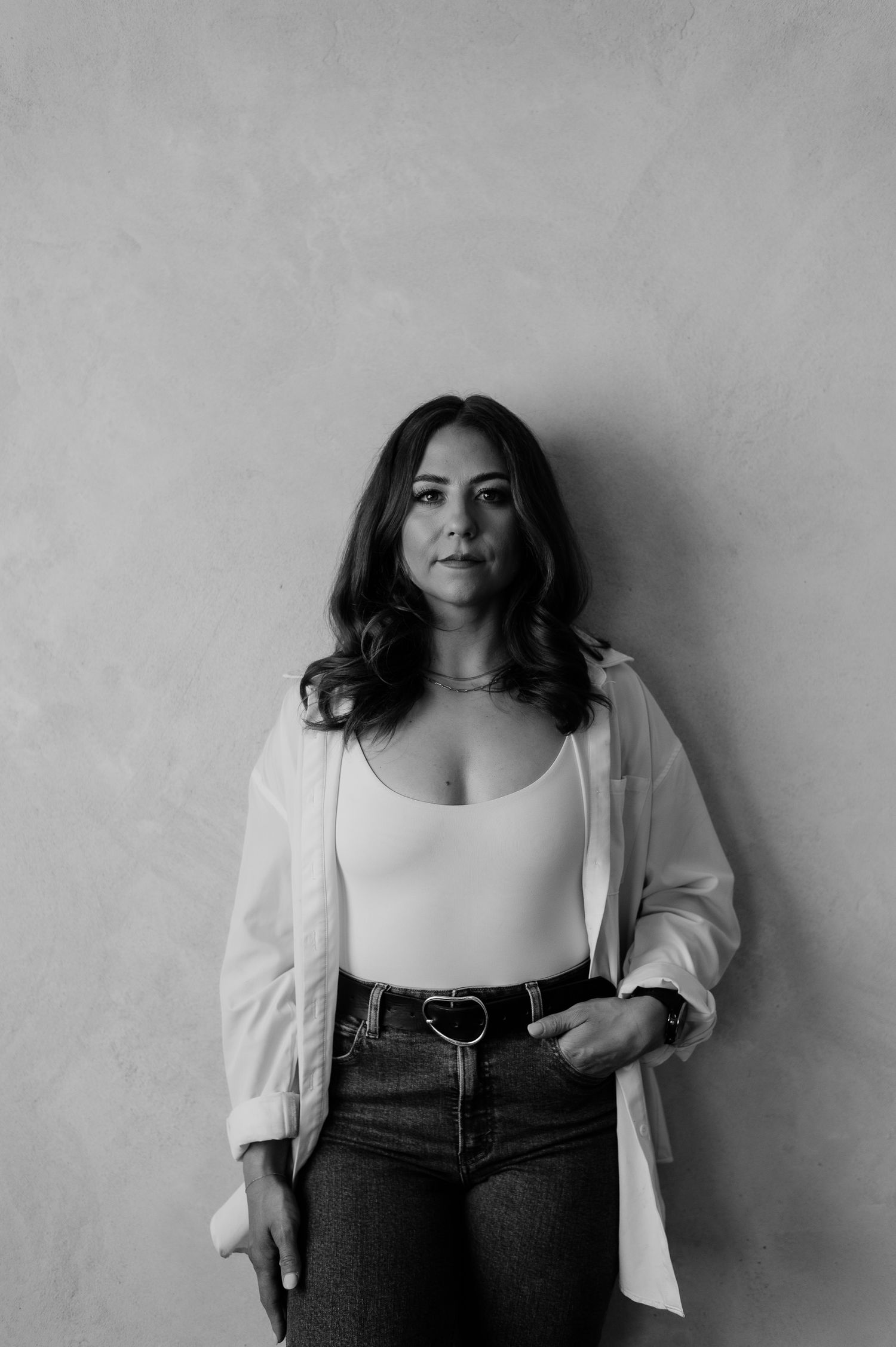 Black and white portrait of a person wearing a white tank top, light blazer and jeans against a plain wall.