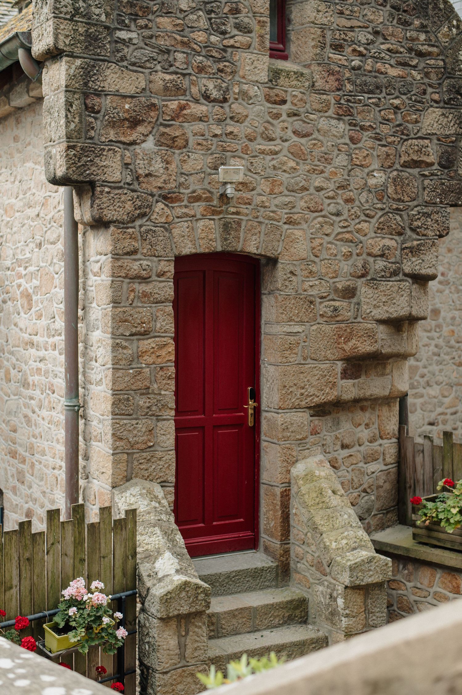 Red door set in a rustic stone building with carved granite archway and steps, surrounded by flower pots.