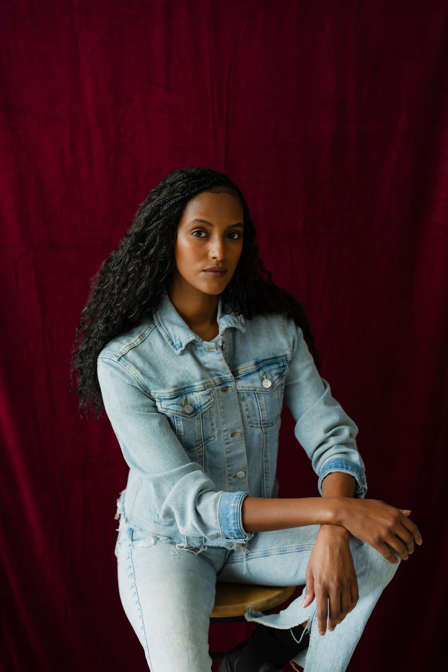 Woman in light blue denim jacket and jeans sitting on stool against red curtain backdrop.