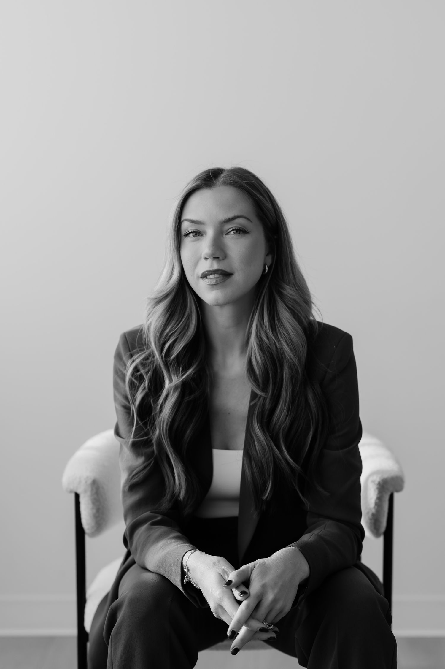 Professional black and white portrait of someone in a dark blazer sitting in a white chair against a light background.