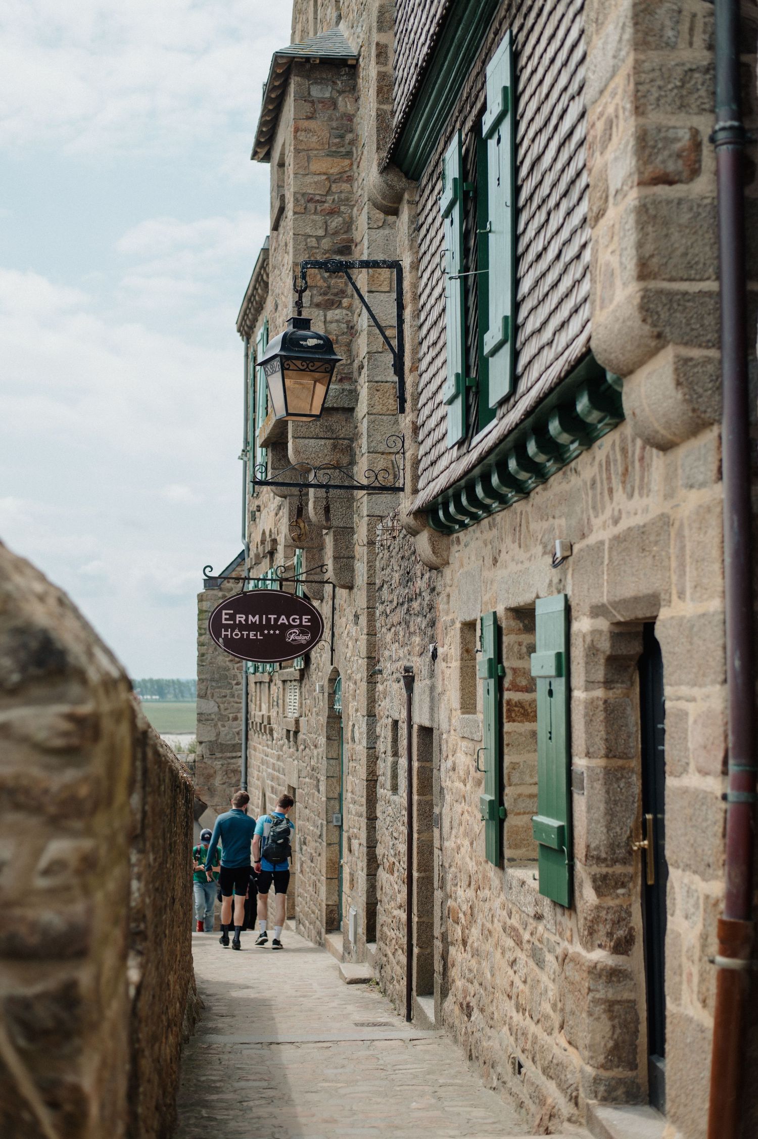 Narrow cobblestone alley at Mont Saint-Michel with stone buildings, green shutters, and Ermitage Hotel sign.