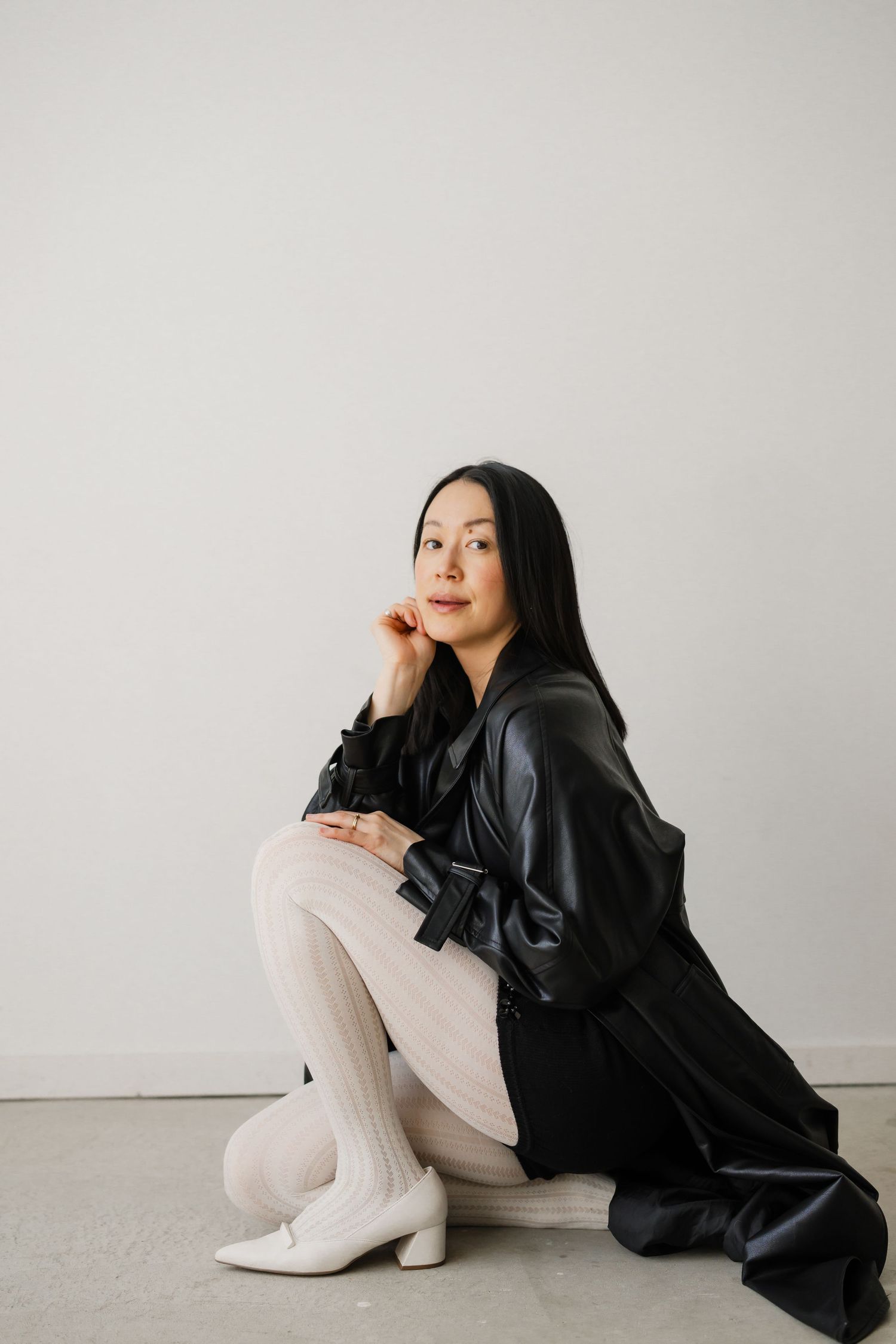 A woman in a black leather jacket and white tights sits gracefully on the floor against a light gray wall.