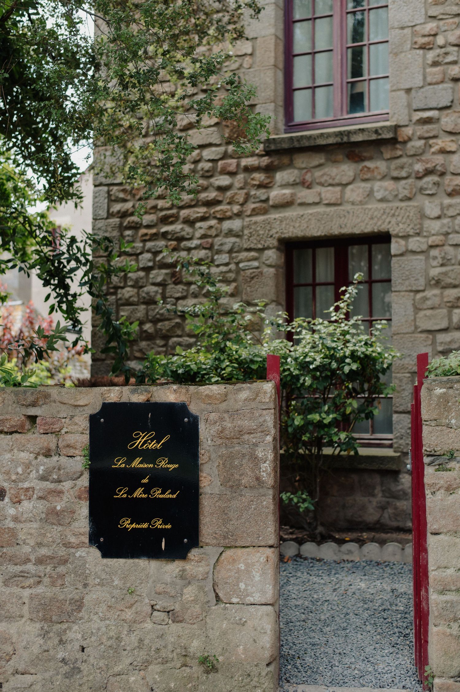 A charming stone hotel in France with a black sign reading Hotel Le Manoir Rouge, framed by lush greenery and red windows.