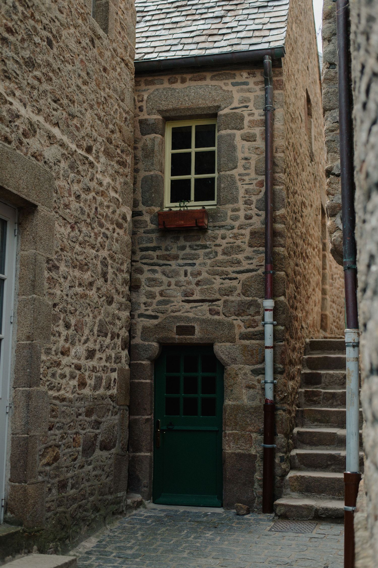 Narrow stone alley with green door and window, exterior staircase, and drainpipe on historic building.