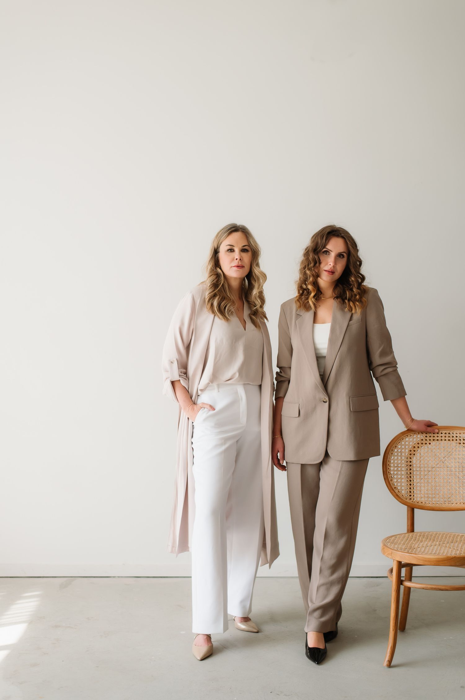 Two models pose in elegant neutral-toned business attire against a white wall with a cane chair.