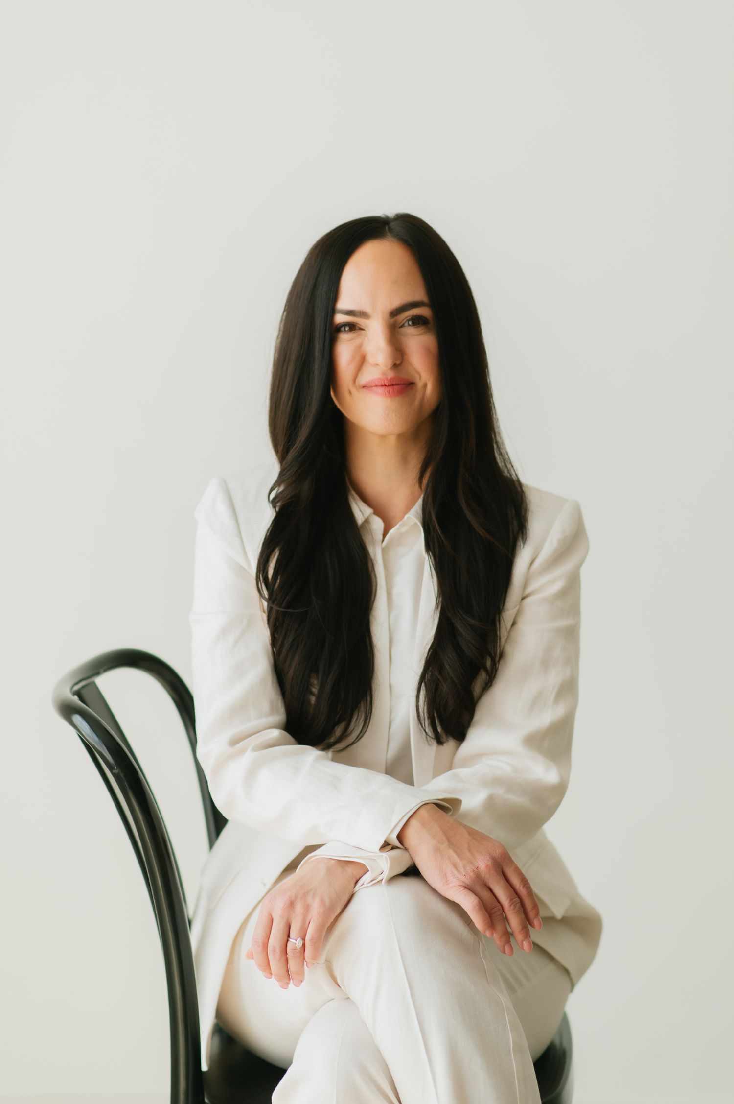 Professional portrait of someone in a crisp white suit sitting confidently on a dark chair against a light background.