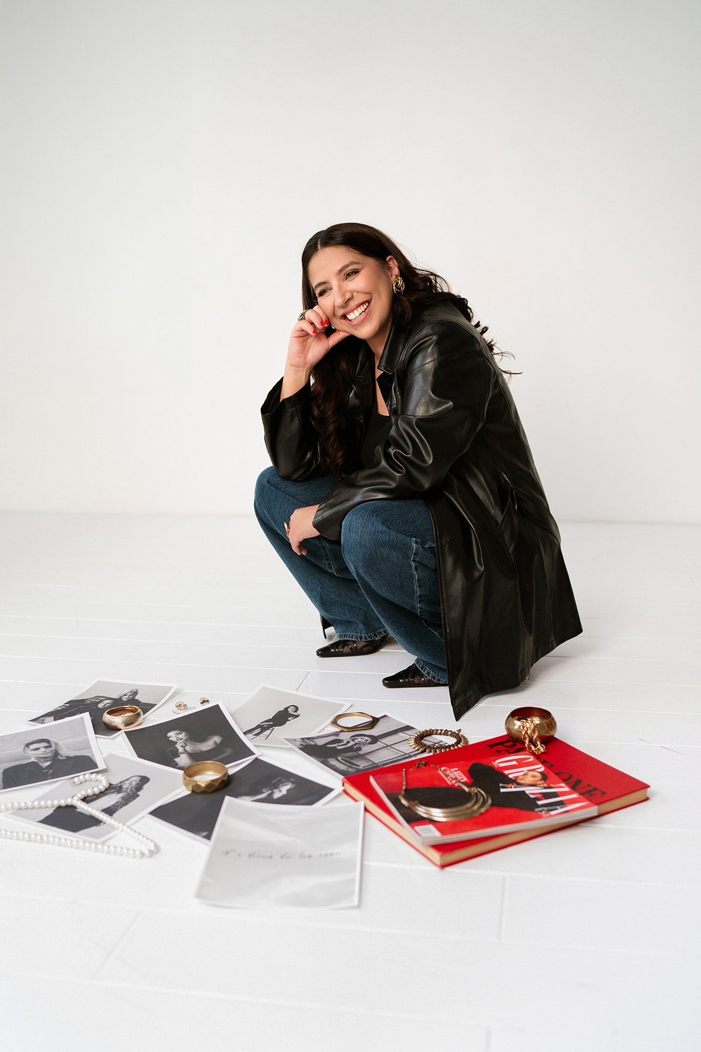 Woman in black leather jacket and jeans crouching beside photos, books, and gold jewelry on white floor.