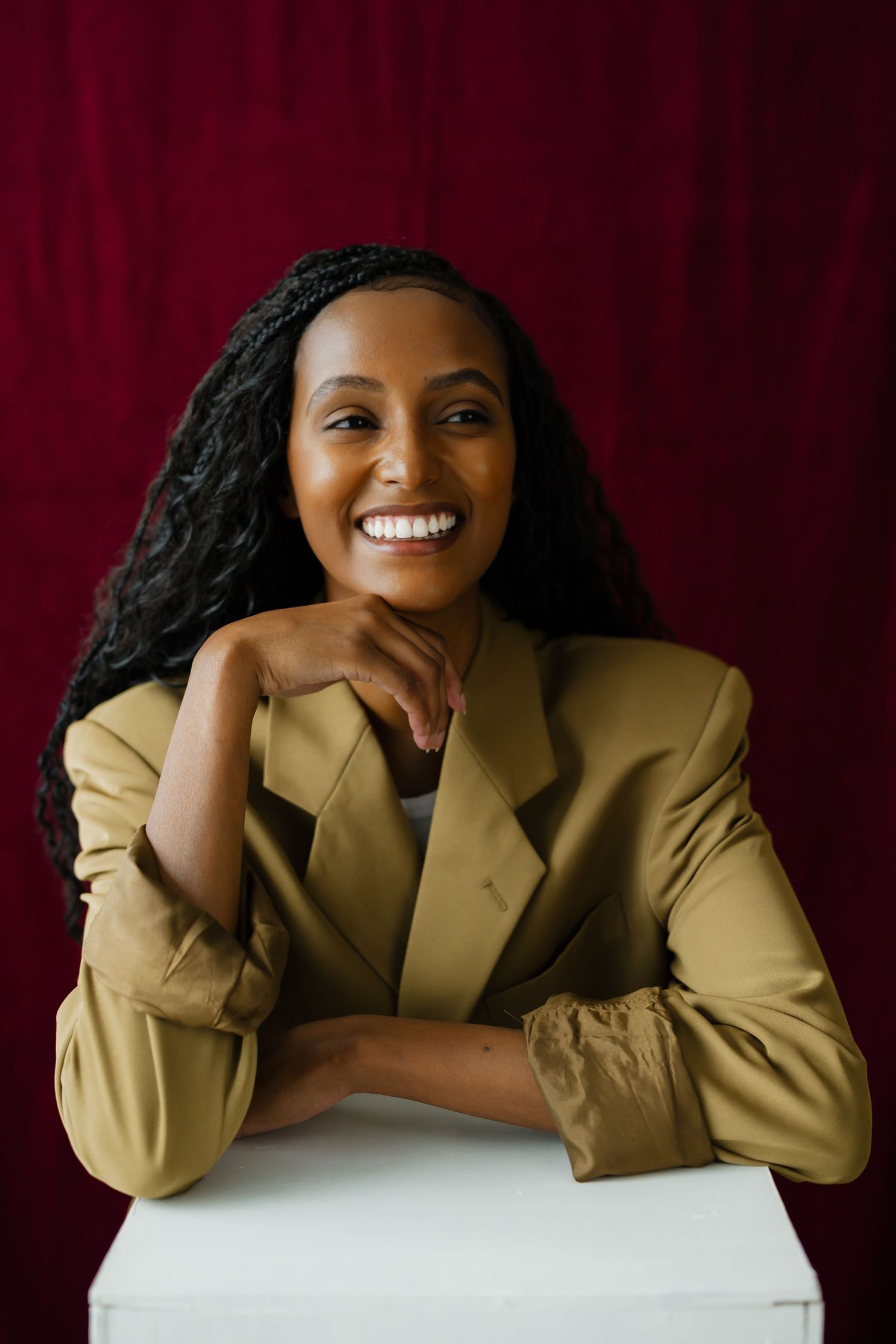 Smiling woman in olive blazer leans on white cube against rich burgundy fabric background.