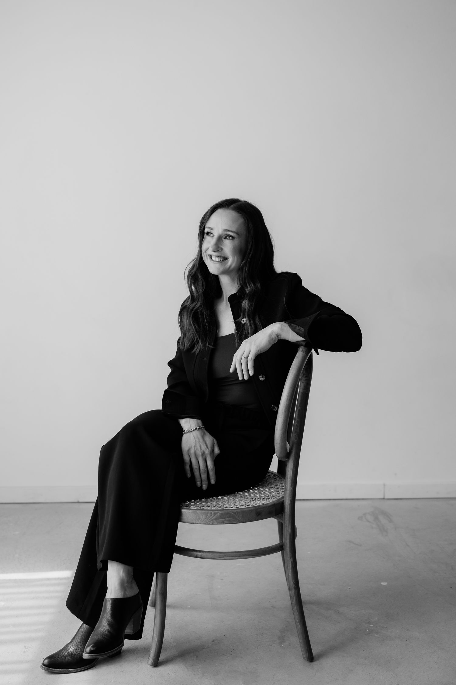 Black and white portrait of a smiling woman in black outfit seated on a wooden chair against a light background.