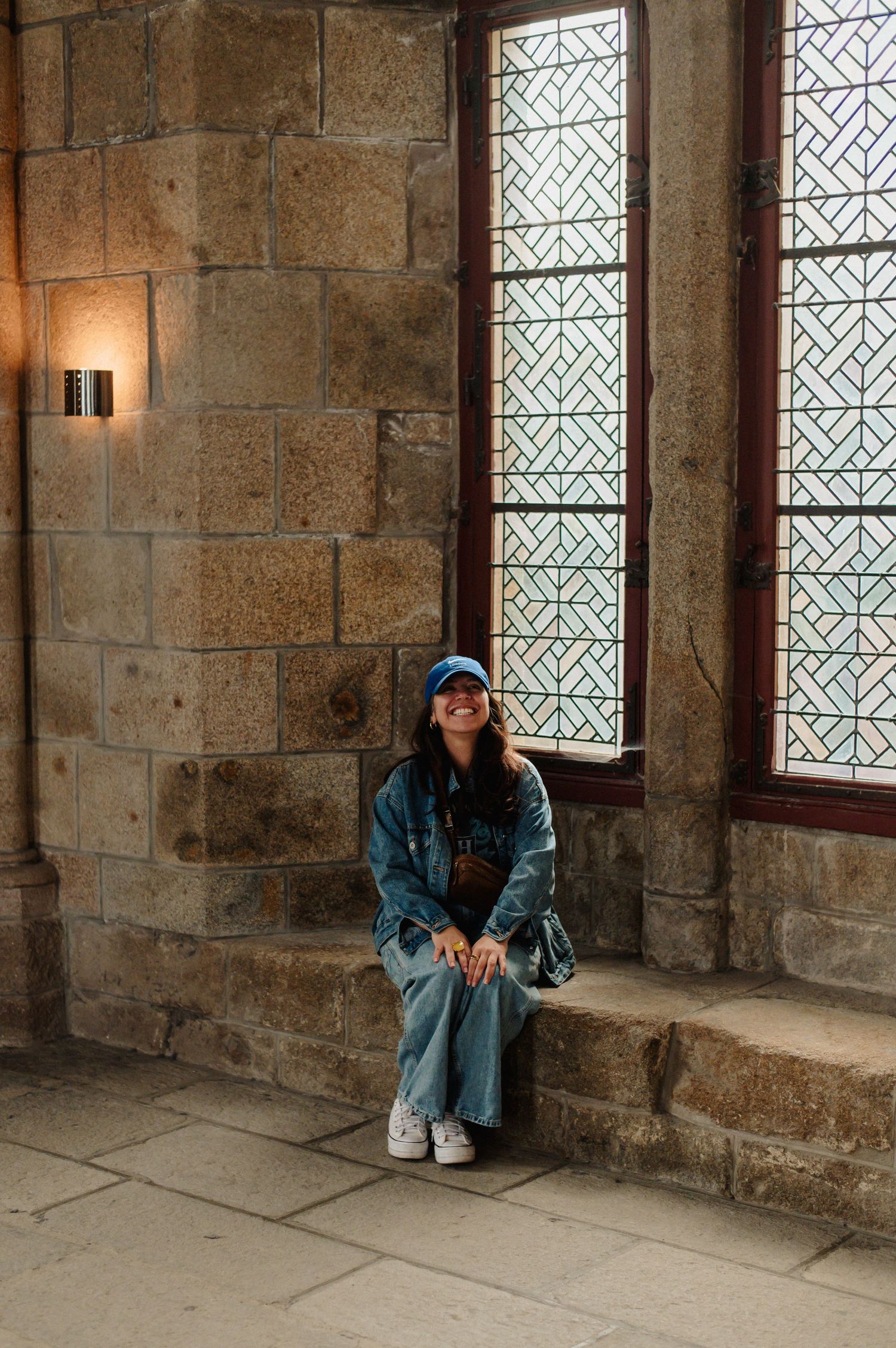 A young woman in denim sits smiling beside ornate latticed windows in Mont Saint-Michel Abbey.