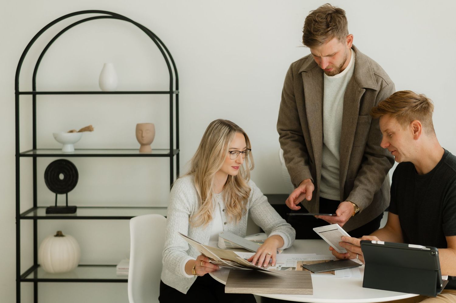 Three colleagues review documents together in a modern office with a minimalist bookshelf in the background.