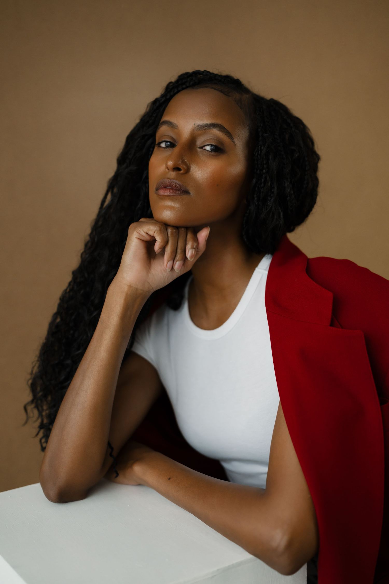 Elegant Black woman with braids in red blazer and white top poses against beige background.